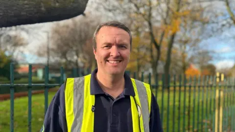 LDRS Michael Brennan smiles as he stands in front of the railings around the park field with tress in the backdrop. He wears a blue top under a sleeveless high-visability yellow jacket.