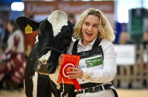 A young woman with blonde hair smiles as she leads her cow inside a ring at the Bath and West Showground near Shepton Mallet