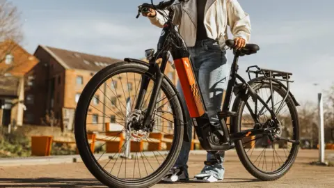 A woman is standing next to a black and orange-coloured e-bike. She is wearing jeans and a cream leather jacket. Her face is not seen.