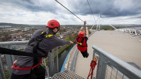 The top of the principality stadium from a platform used to launch the zip liner. A man on the left of the photo is strapped on with a black strap. His arm is outstretched towards a woman who has just launched. He is hanging on to a handle next to the zip line and roller. She is strapped in with a shoulder harness visible. The roof of the stadium and the city beyond are in the background. 