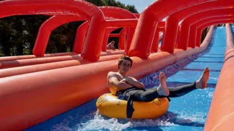 A man with his arms crossed is riding a yellow inflatable tube down a wide water slide filled with shallow water, surrounded by large red inflatable arches that create a tunnel-like structure over the slide.