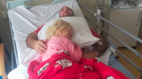 Emma Alderman Noa-Rose sleeping on a pillow on top of Scott in a hospital bed. Noa-Rose wears pink pyjamas and has a red blanket on op of her. Scott has his arm around her and stares up to the ceiling with a worried look.