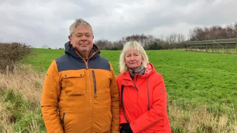 BBC/ Olivia Richwald Two people stand in a grassy field on an overcast day, one in a yellow and navy jacket and the other in a red jacket, with a row of solar panels and a line of trees visible in the background.
