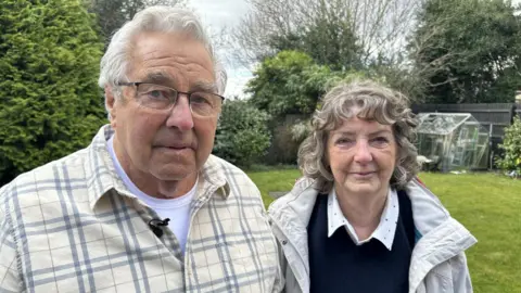 John Baker, with his wife Janice in their back garden. There are trees and grass behind them. She is wearing a blue jumper and a shirt and he is wearing a yellow  and grey shirt. They are both of retirement age and looking into the camera. 