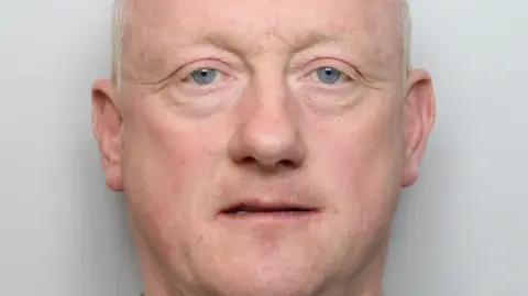 Mugshot of Christopher, against a grey backdrop. He has short white hair, blue eyes and thin eyebrows.