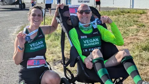 David and Alice hold up medals after a run at Solent Airport. Alice is crouched alongside David's wheelchair. Both wear running clothes and are smiling at the camera.
