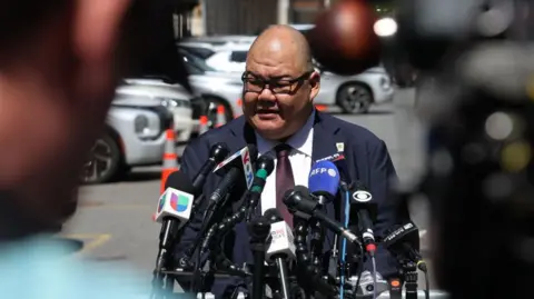 Getty Images Steven Cheung speaking into a lot of microphones at a press conference in a carpark, with several white cars parked behind him.