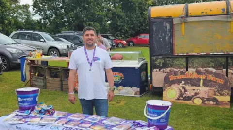 Trevor Bygate A smiling Trevor behind a stand with Papyrus leaflets on it, in a field with signs that read "Bramblebee Farm"