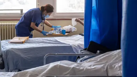 PA Media A hospital ward with several beds separated by blue privacy curtains. A healthcare worker in blue scrubs and a face mask leans over a bedside table while attending to a patient beside a bed. Large windows provide daylight, and medical equipment and bedding are visible throughout the room.