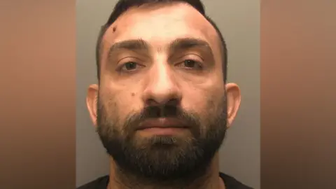 Police mug shot of Hristo Iliev. He has short dark hair and a beard and is looking directly at the camera.