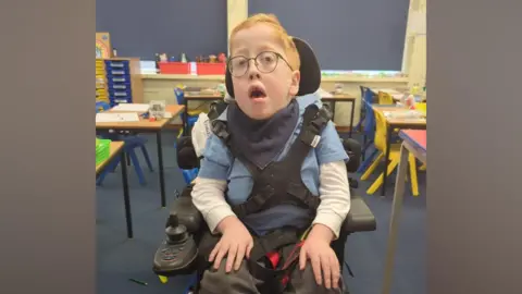 Ezra, sat in an electric wheelchair and looking at the camera. He is within a colourful classroom with small chairs and desks.