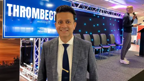 A man in a grey suit, white shirt and blue tie, stands to the side of a stage, smiling