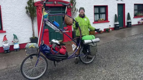A man is standing beside a large bicycle. The seat and handlebars are taller than his head. The bicycle is painted purple and has baskets and bags attached on the front and back. The man is wearing a green coat and hat. 