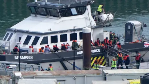 PA Media A Border Force vessel is moored alongside a dock in Dover on 23 July 2026 as a group of people wearing orange life jackets disembark. Several officials in high-visibility clothing stand on the quayside and gangways, with harbour buildings and water visible in the foreground and background.