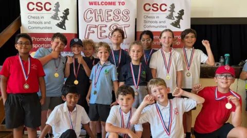 Chess in Schools A group of about a dozen primary school children are posing in front of large posters saying 'CSC Chess in Schools and Communities'. They all have medals around their necks and some have got their thumbs up or arms in the air.