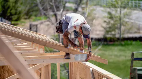 Getty Images Drone View of a house being built by carpenters in the US. A builder is hammering in a nail on a roof frame.