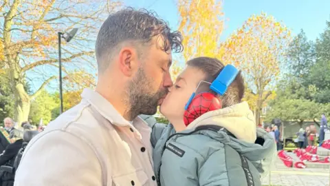 Chris Jefferson A man kisses a young boy outside. The man has dark brown short hair, a cropped beard and moustache. He is wearing a camel-coloured jacket. The boy has dark brown hair and is wearing a pale green-coloured jacket, grey trousers and head phones. 