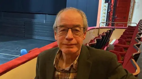 A man with short grey hair and glasses smiles at the camera. He is sitting in a theatre auditorium, wearing a checked shirt and green blazer.