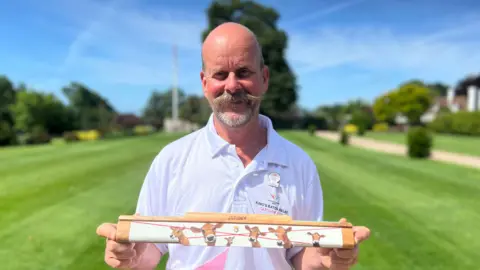 Person standing outdoors on a sunlit lawn holds a wooden relay baton decorated with painted cow illustrations; the person wears a white “King’s Baton Relay Glasgow 2026” polo shirt, with trees, a path, and buildings blurred in the background under a blue sky.