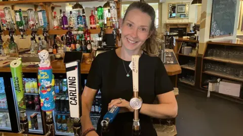 BBC/Kate Baldock A woman standing behind the bar in a pub pulling a pint. She is wearing a black t-shirt and smiling. She has light brown hair which is tied back in a ponytail and is wearing a necklace with a teal heart pendant. Her head is tilted to the right. 