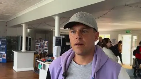 A man wearing a grey baseball cap and a purple hoodie. He is stood in a conference room looking at the camera