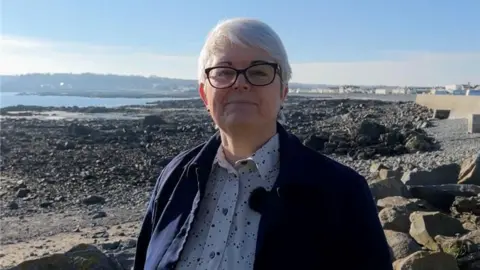 Hannah Beacom - a woman wearing a spotty white shirt with a navy overshirt standing on a rocky beach.