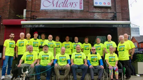 Ova the Hills A group of 20 men and women, who are wearing matching yellow T-shirts with the walking route on them, have gathered together for a photograph. 
