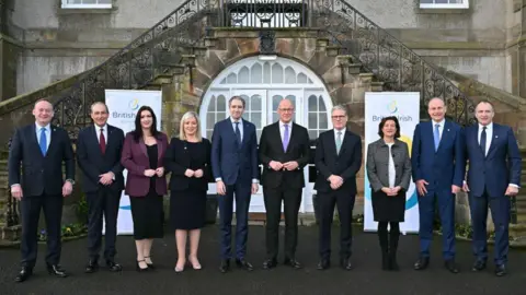 PA Media A family-style photograph of the 10 members of the British and Irish council standing outside a building with a white arched door. The seven men in the photo are all wearing dark suits, they include John Swinney who is bald with glasses and wearing a purple tie and Keir Starmer who has grey hair and is wearing a green tie. 