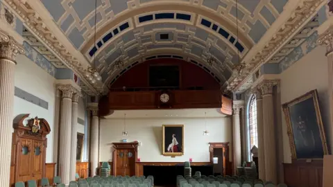 Ornate geometric pale blue and cream curved ceiling in room with pale green seats. Wooden panelling and a clock can be seen on one wall and ornate a wooden doors. A portrait of the late Queen Elizabeth hangs on one all with other artworks. 