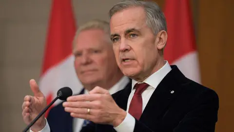 Prime Minister Mark Carney speaking at a podium. Behind him is a row of Canadian flags. He is wearing a black suit, a white collared shirt underneath and a burgundy tie, with a small Order of Canada pin on the collar of his suit jacket. He has short grey hair. 