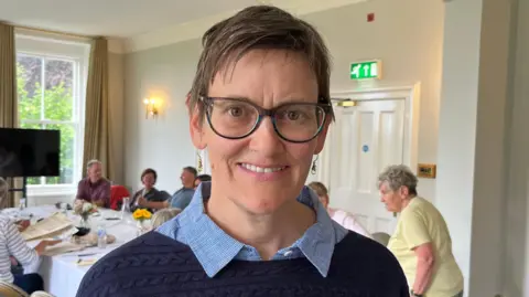 A woman with short brown hair and glasses with people sat around a table in the background