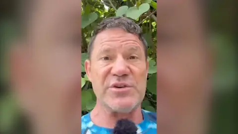 Steve Backshall stands in front of some foliage. He is speaking into a mic and wearing a mottled blue T-shirt.