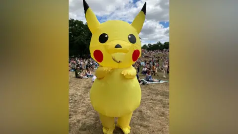 Ollie Ward/BBC A person dressed in a large yellow Pikachu Pokemon costume in a park. Crowds of people are behind them.
