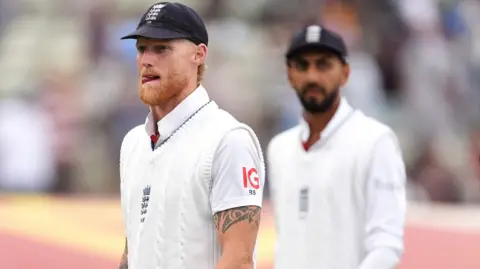 Ben Stokes, wearing a blue cap and white sweater, looks frustrated, left, with Shoaib Bashir in the background