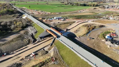 HS2 A large area of construction surrounded by green fields. In the centre is a newly built bridge carrying the base of a new railway line over a track below. It is surrounded by large machinery, piles of earth and temporary cabins stretching into the distance.