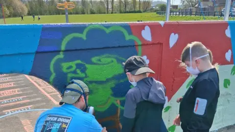 Open Door Art Three children in tracksuits wear protective masks and help a professional artist spray paint the mural