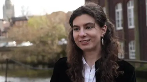 Shaun Whitmore/BBC Alessia Oancea standing next to the river in Norwich. She has brunette hair and is wearing a shirt with a black jacket over the top. She is looking towards the left of the camera and is smiling.