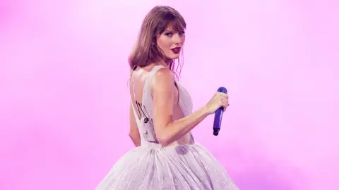 Getty Images Taylor Swift, in front of a pink background. She is wearing a glittery dress and holding a microphone