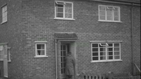A man opening the front door of a semi-detached brick house.