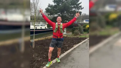 A middle-aged man wears a red training top, dark grey shorts, red socks and green trainers, as he stands in a park with both hands raised in triumph above his head.