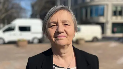 Deputy Mary Le Hegarat is standing in a paved square in front of a road, with an office building behind it. She is wearing a black blazer and is looking at the camera.