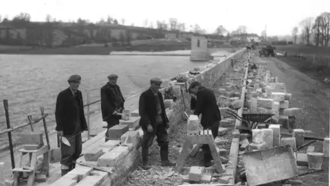 A black and white photo of four men working on some brick work by the edge of the water. They have flat-caps on and are dressed smartly in wellies and dark clothing. You can see the wall of the edge of Chew Valley gradually being built with the water tower in the background.