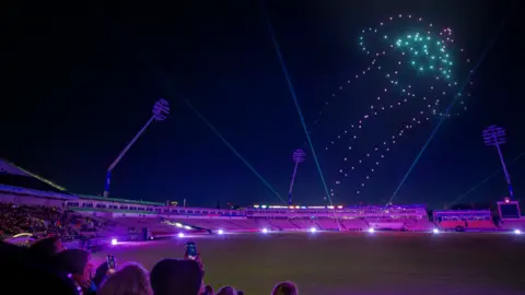 Yuup Drones create the shape of a jelly fish in the night sky. The ground is lit up in pink and purple and people are sitting watching the display.