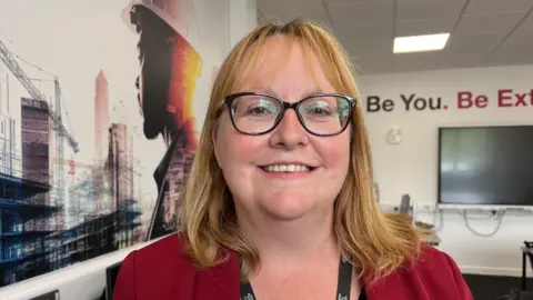  Woman with glasses and blond hair smiles at the camera, she is standing in classroom with a builder artwork behind her