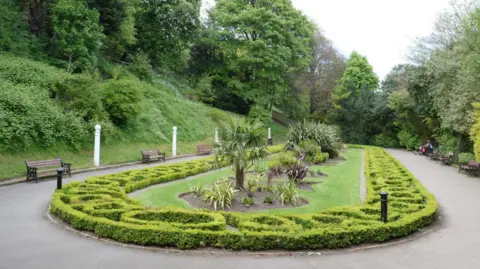 One of the ornate displays with the garden. It features a grassed area surrounded by planting and some benches. The landscaped area has the same shape as a race track, with curved ends and long straight sides. It is bordered with intricately shaped, very low hedgerows. A neat lawn sits inside the hedge-line and has a series of small trees and bushes in a straight line down the middle. To the left, the gardens rise in a steep slope.