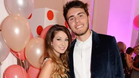 A woman and a man stood side by side in front of balloons. They are both smiling and looking directly at the camera. 