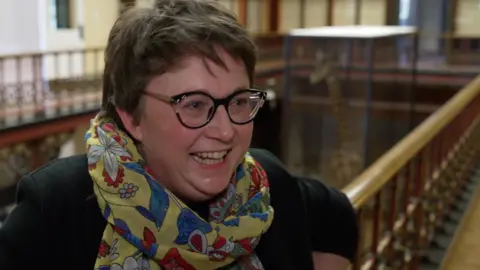 John Fairhall/BBC Woman in bright colourful scarf smiles off camera and is standing on a mezzanine of a museum