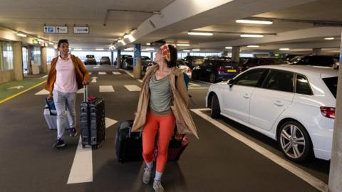 A man and woman drag multiple suitcases behind them through an airport carpark