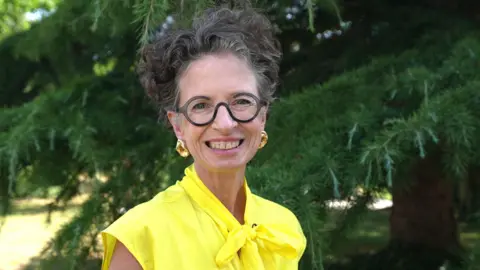 Liz smiles at the camera. She has grey curly short hair and wears glasses with round frames. She wears a bright yellow top and gold statement earrings, and stands in front of a large tree. 