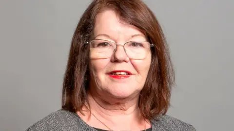 UK Parliament Head and shoulders image of Kate Hollern against a grey background. She has shoulder length brown hair and is wearing glasses and smiling